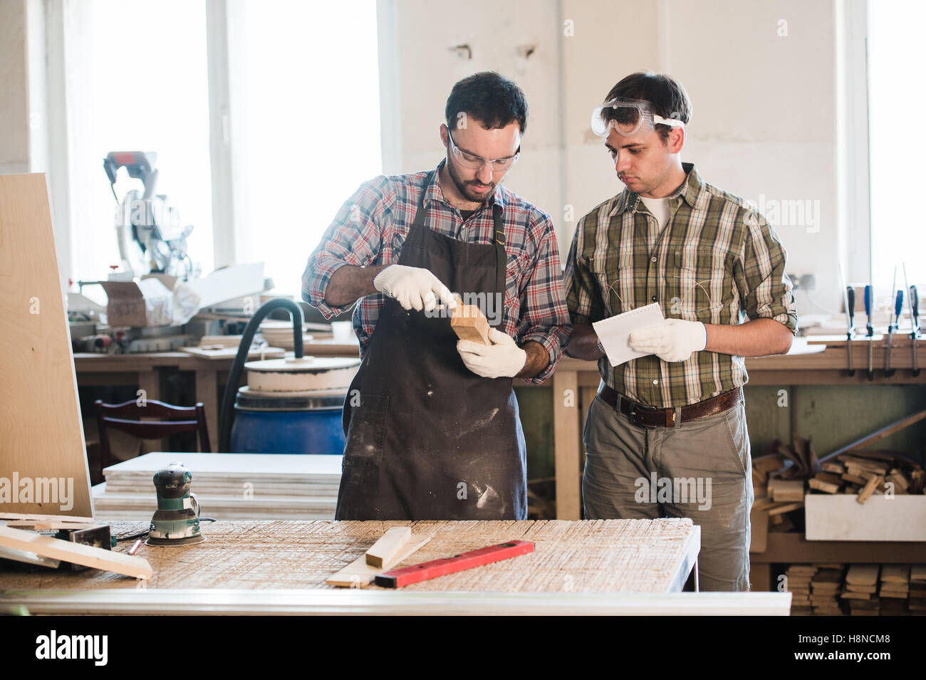 Happy male carpenter showing something to coworker at his notebook papers in workshop Stock Photo