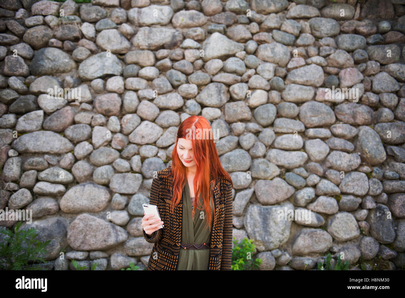 Redhaired woman standing against stone wall and using phone Stock Photo
