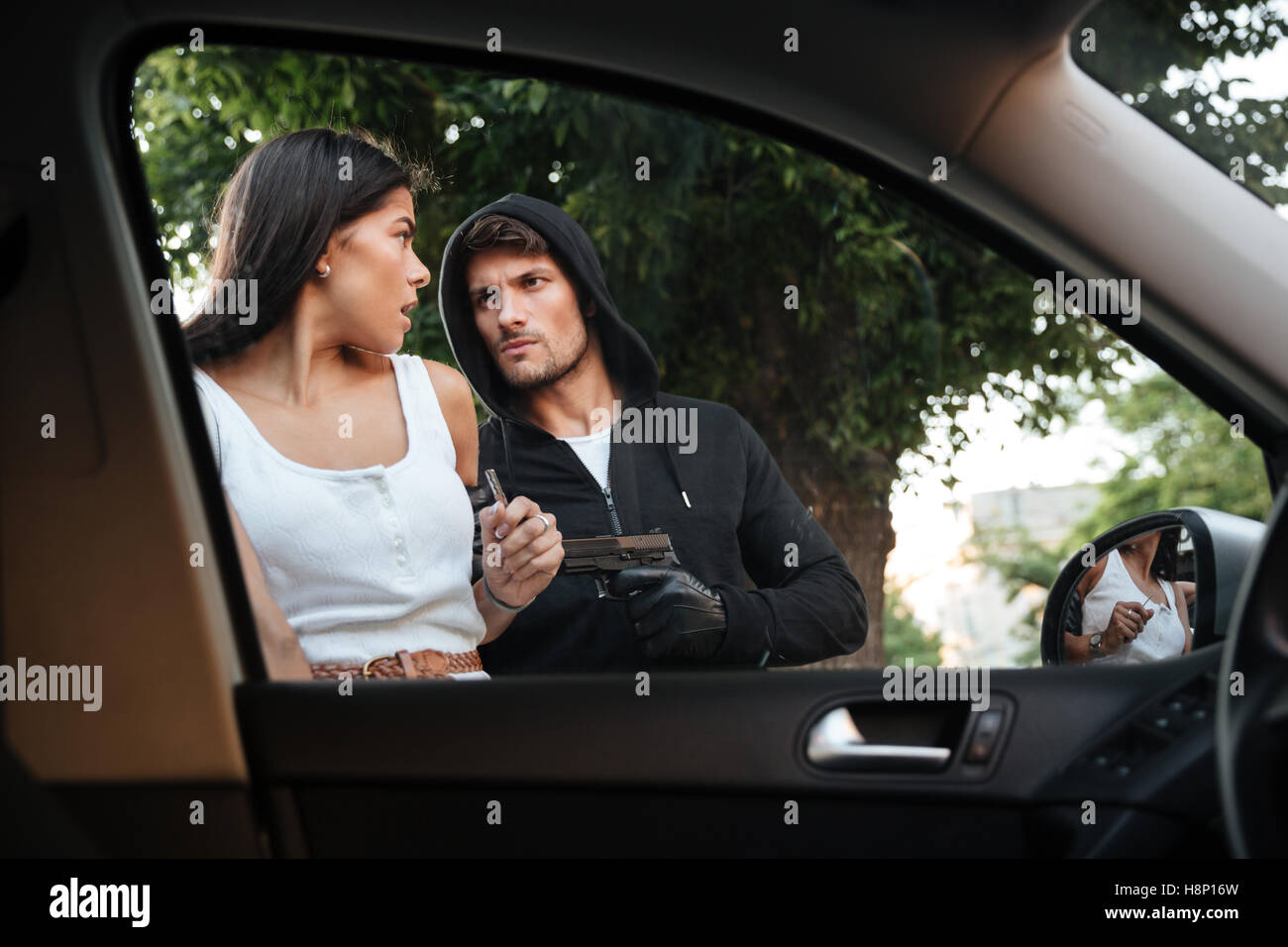 Dangerous criminal man with gun stealing car of scared young woman on outdoor parking Stock Photo