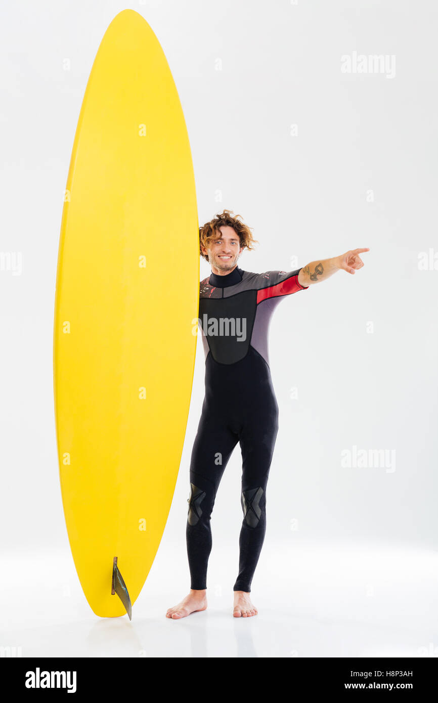 Cheerful young surfer holding surfboard and pointing finger away isolated on the white background Stock Photo