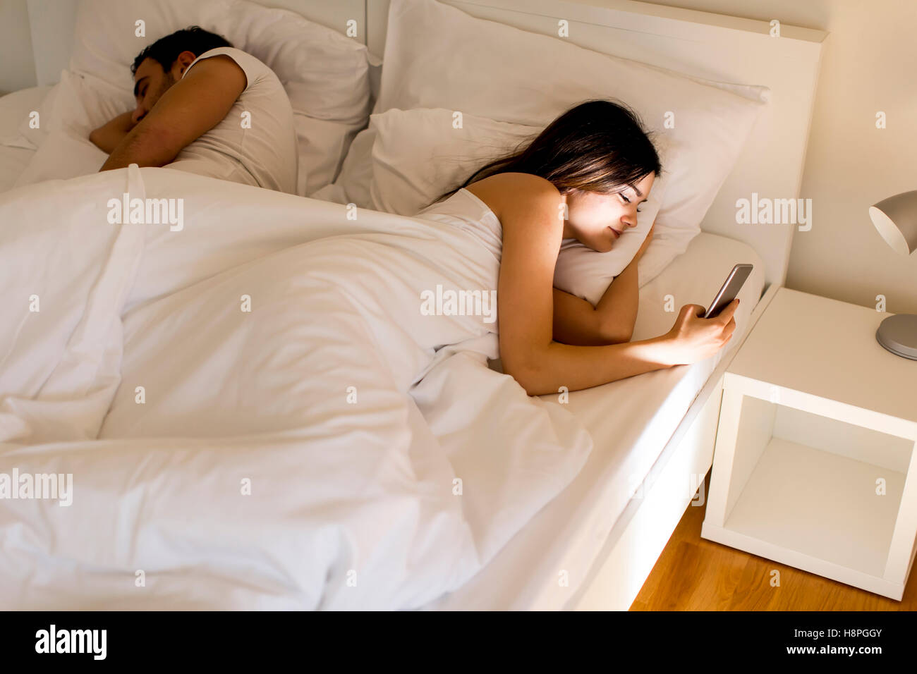Young couple in bed. Man sleeps while a young woman looking phone at night Stock Photo