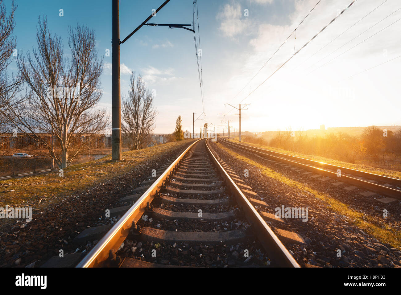 Railroad against beautiful sky at sunset. Industrial landscape with railway station, colorful blue sky, trees and grass, sun Stock Photo
