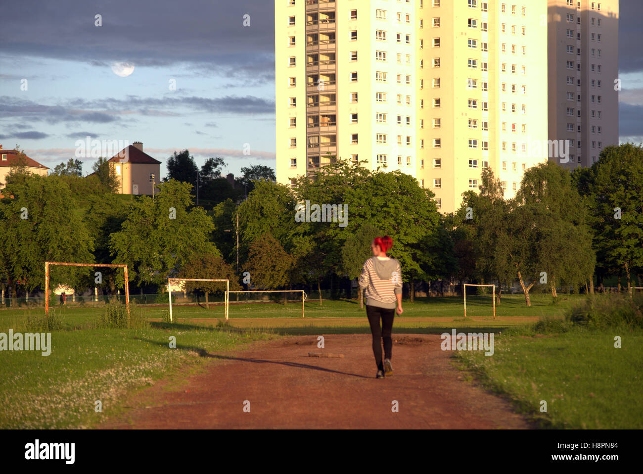 Red haired girl walking along   lincoln avenue football pitches with the sun and moon high flats or skyscrapers Stock Photo