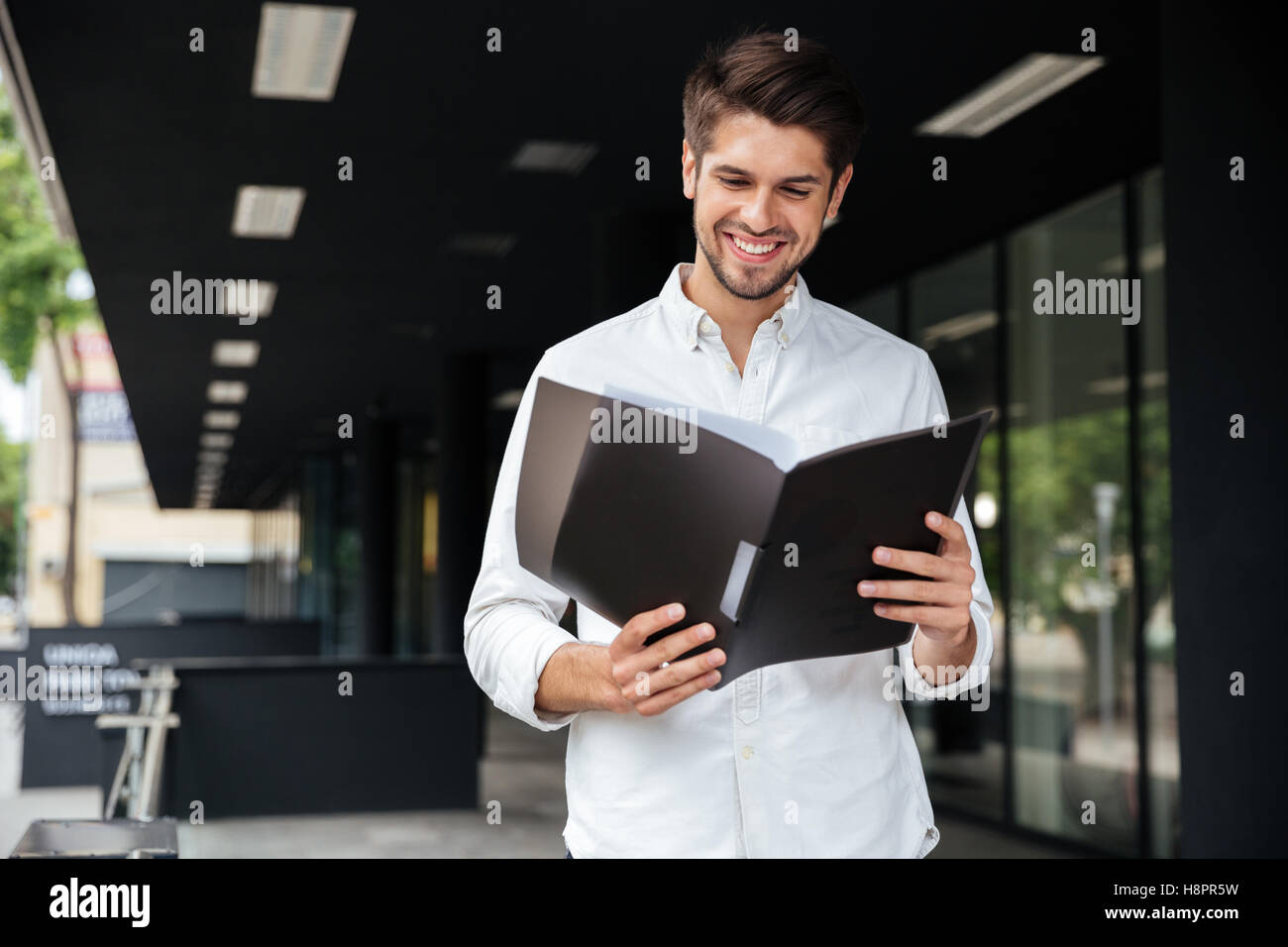 Happy handsome young businessman standing and looking through documents in folder Stock Photo