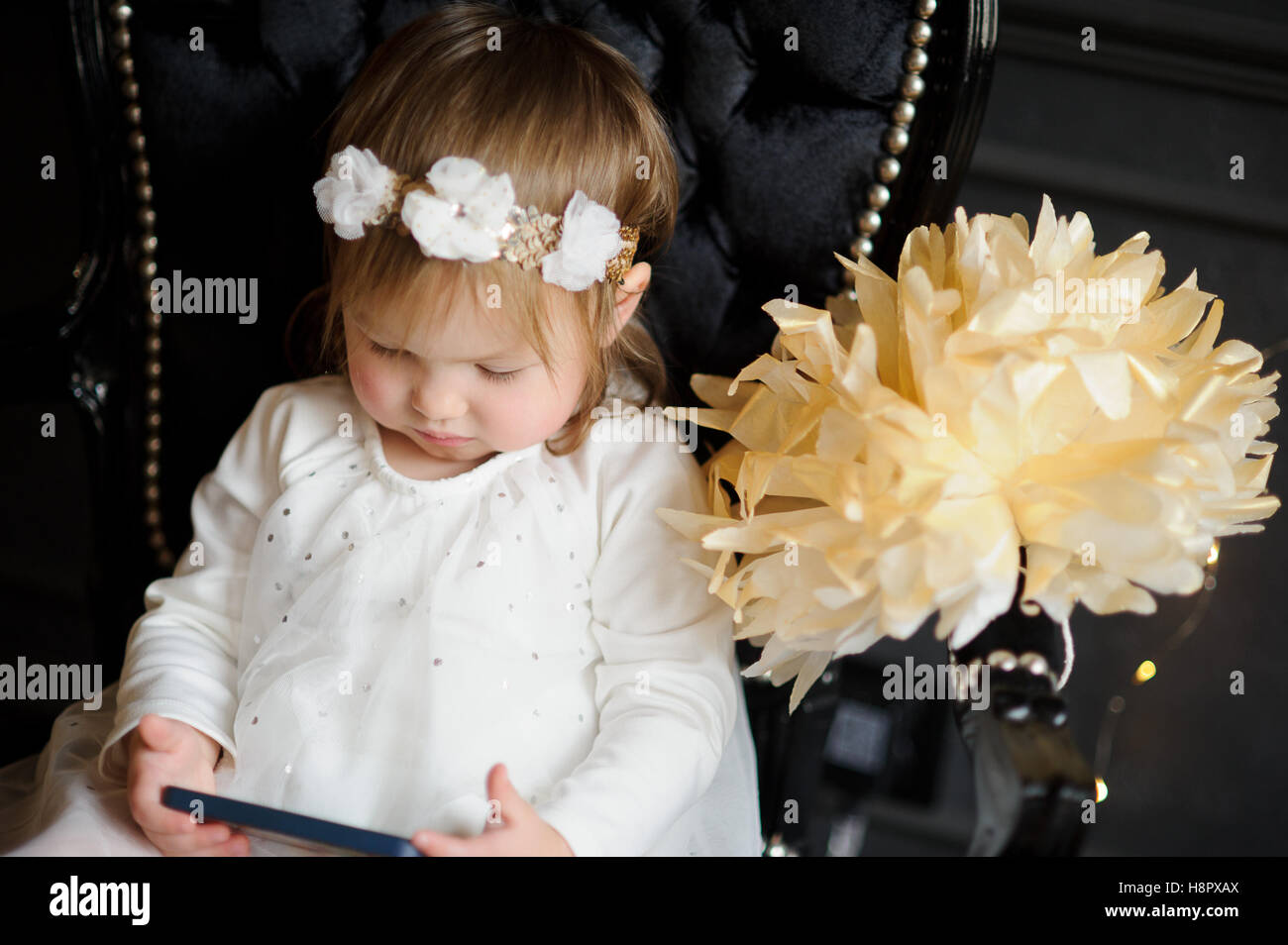 Bonny ruddy toddler sitting in the chair and enthusiastically looking at smartphone screen. In white dress, with a wreath on his Stock Photo