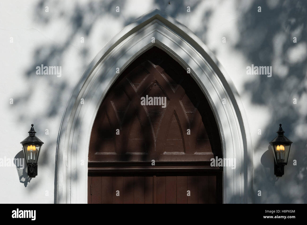 An arched old brown church door flanked by lamps, sprinkled with shadows from a tree. Stock Photo