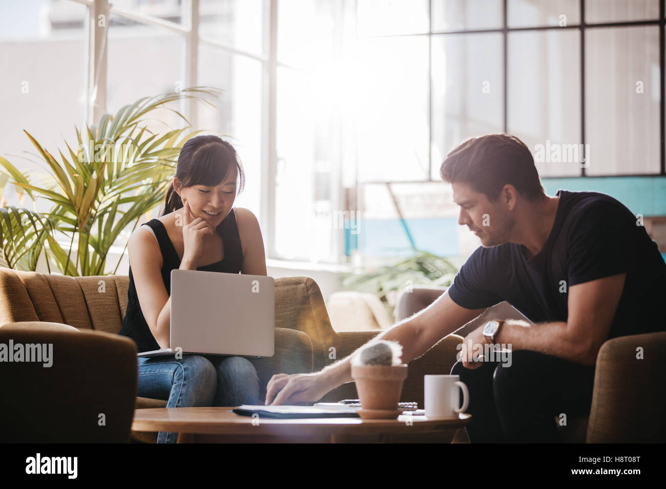 Shot of two people sitting in office lobby and working on laptop. Asian woman and caucasian man working together in modern offic Stock Photo