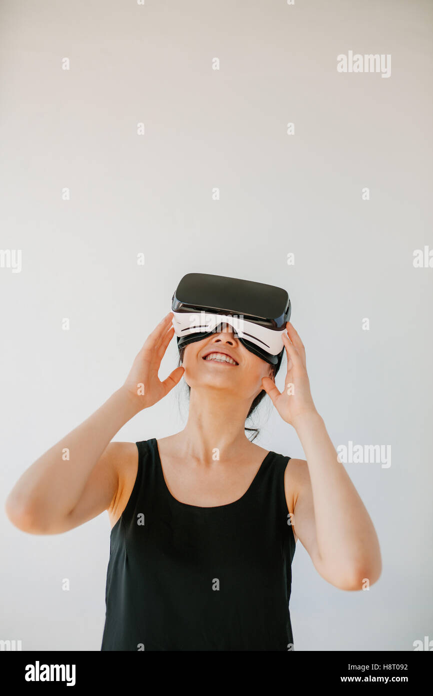 Vertical shot of young woman using the virtual reality headset against grey background. Happy female model wearing VR goggles. Stock Photo