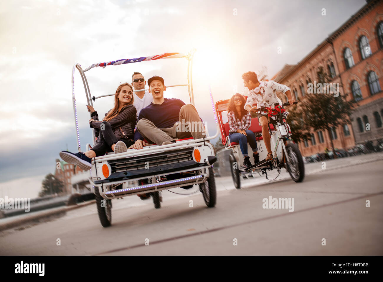 Happy young friends going on tricycle ride through the city. Teenagers sitting on tricycles on road. Stock Photo
