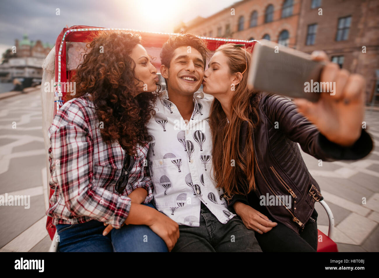 Women kissing man and taking selfie on tricycle. Group of friends having fun on holidays. Stock Photo