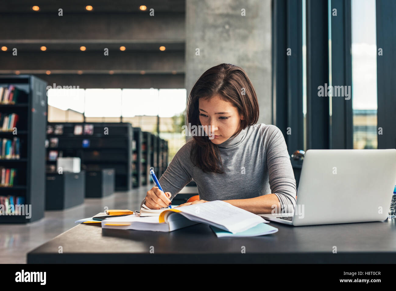 Female student taking notes from a book at library. Young asian woman sitting at table doing assignments in college library. Stock Photo