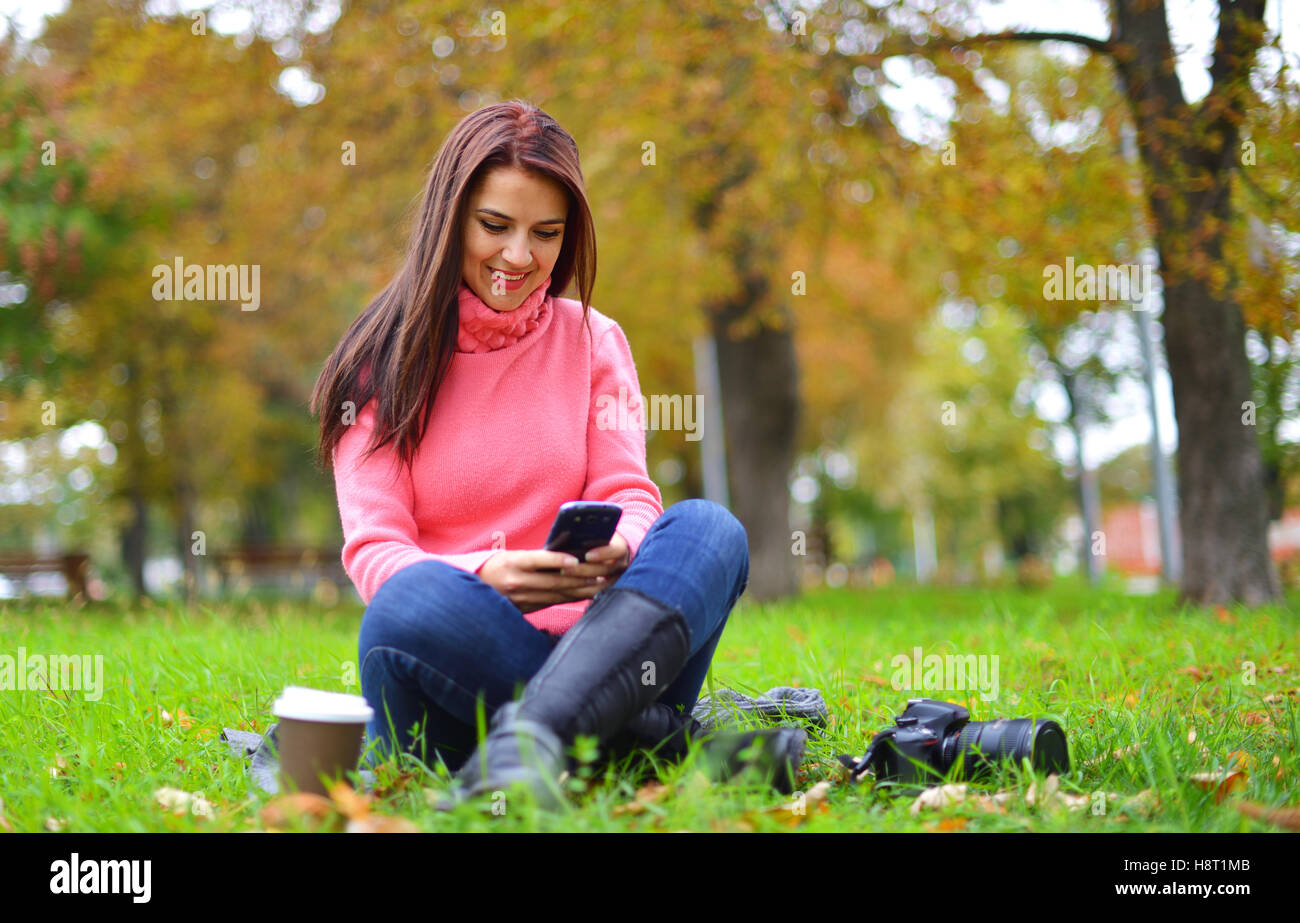 Young fashionable teenage girl with smartphone, camera and takeaway coffee in park in autumn sitting and smiling Stock Photo