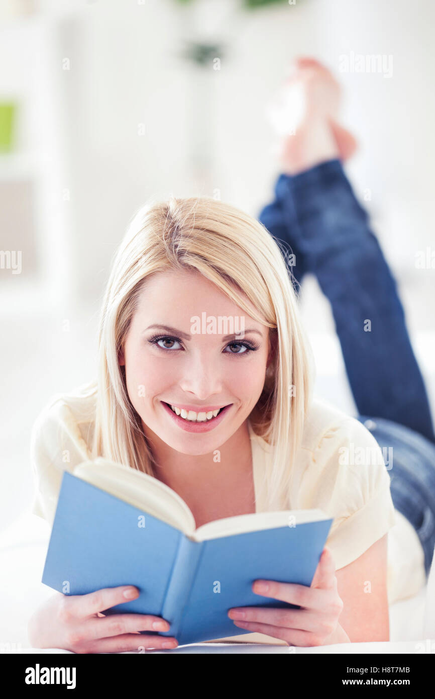 Beautiful young Caucasian woman lying on sofa and reading a book in living room. Looking at camera. Stock Photo