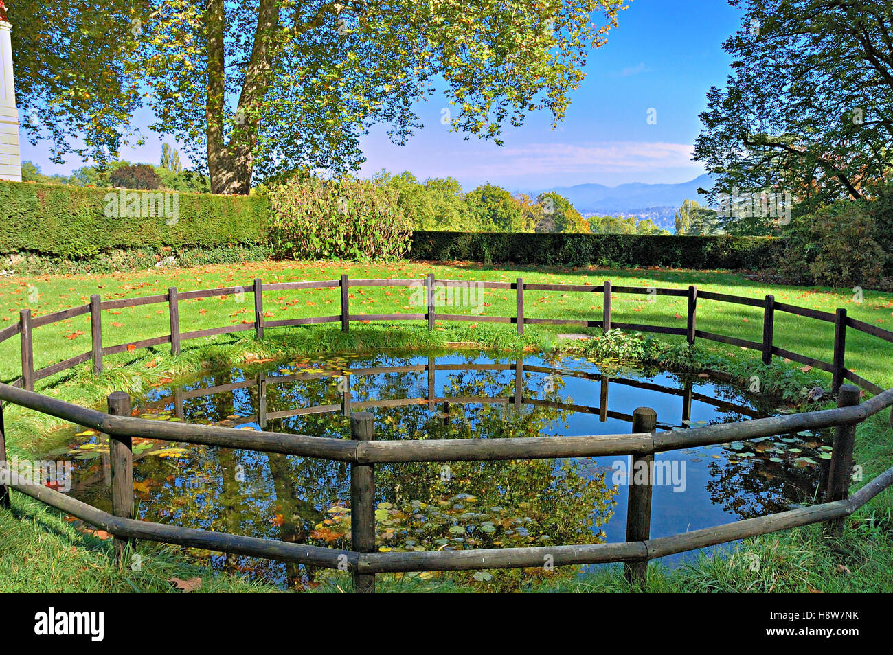 Pond circled by old wooden fence in a garden with mountains in the distant background. Stock Photo