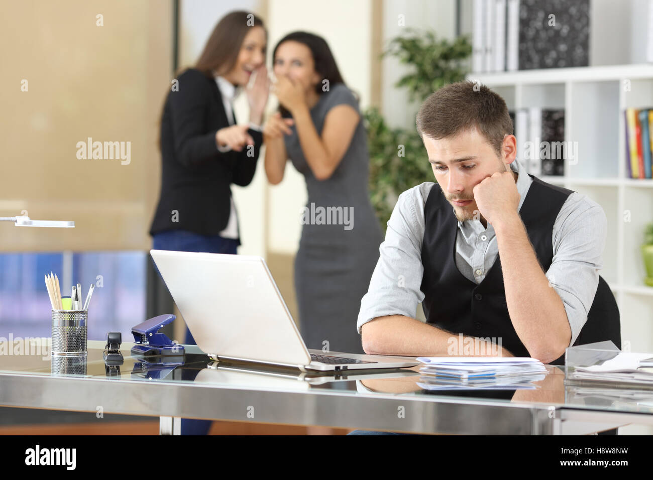 Two coworkers bullying a worried colleague that is sitting in his workplace at office Stock Photo