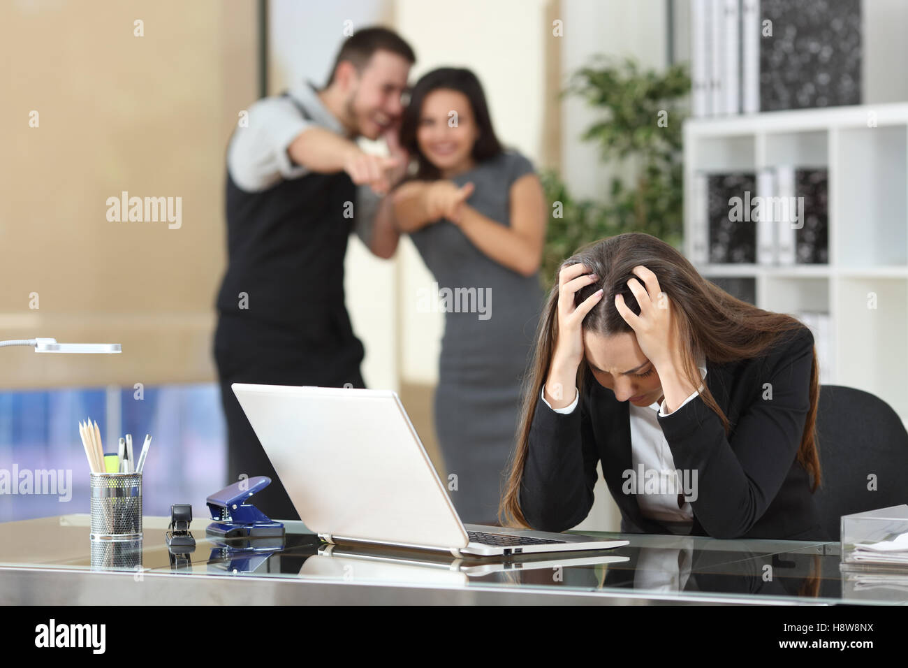 Two businesspeople bullying a sad colleague that is sitting in her workplace at office Stock Photo