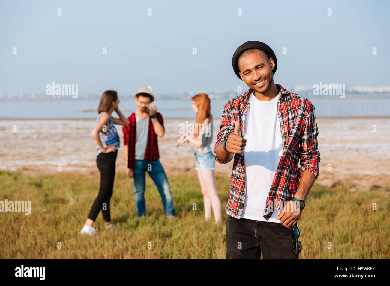 Happy african young man standing and showing thumbs up while his friends talking outdoors Stock Photo