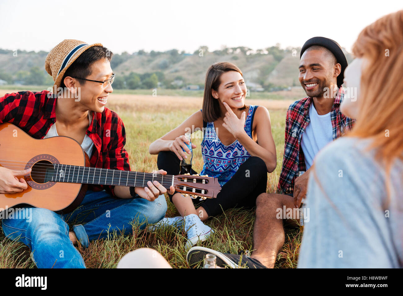 Group of smiling young friends drinking beer and playing guitar outdoors Stock Photo