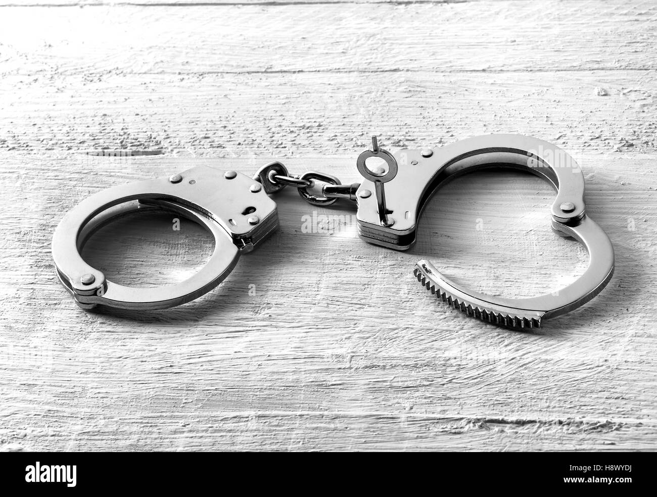 Pair of handcuffs with one side unlocked and the key in the lock lying on a textured white wooden board with foreground copy spa Stock Photo