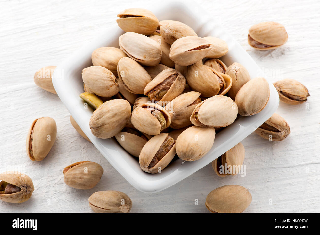Roasted plain pistachio nuts in a square bowl spilling over onto a white textured wooden board below, high angle view Stock Photo