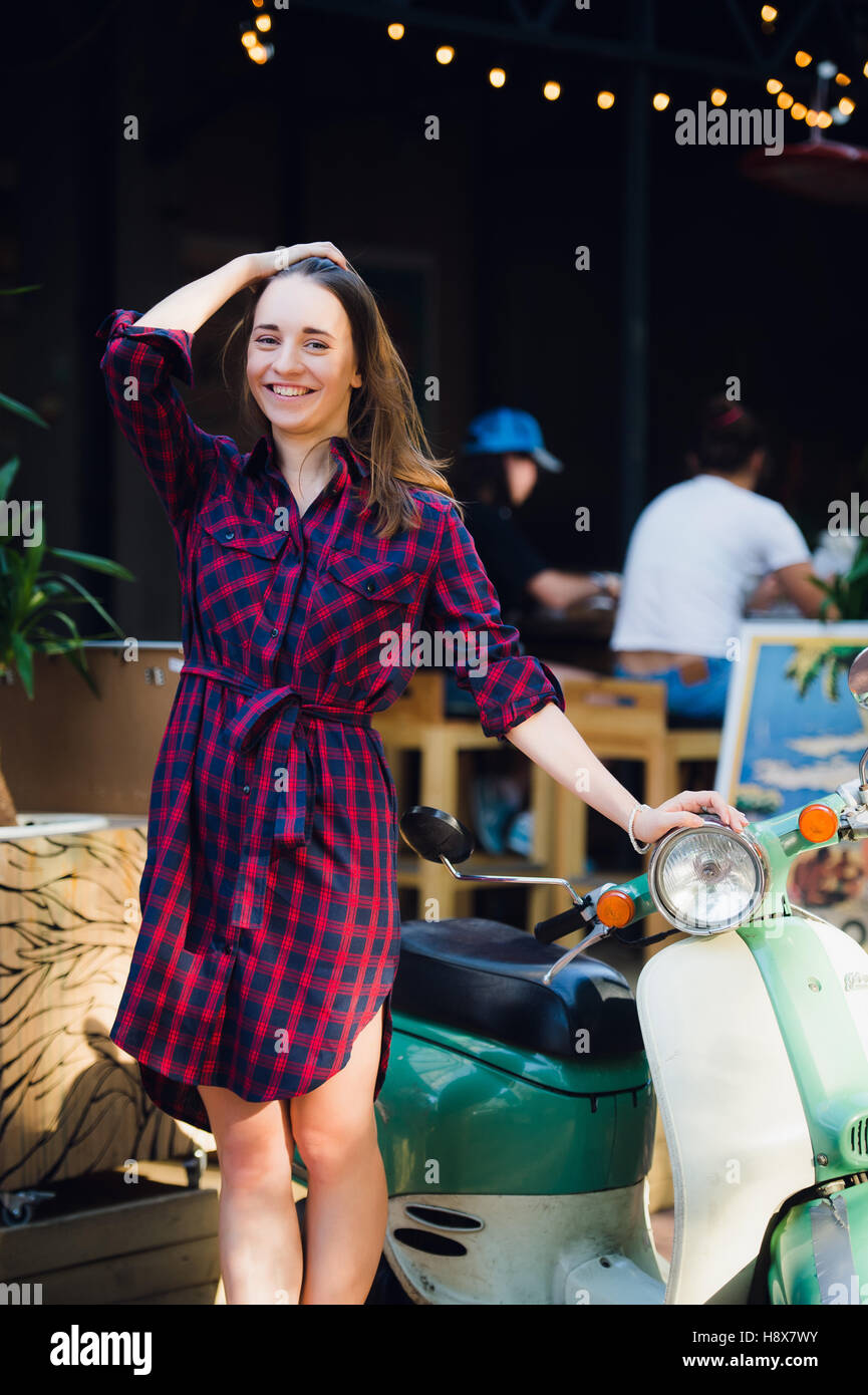 Urban fashion. Outdoor portrait of pretty young woman wearing checkered dress standing near scooter at park outdoors Stock Photo