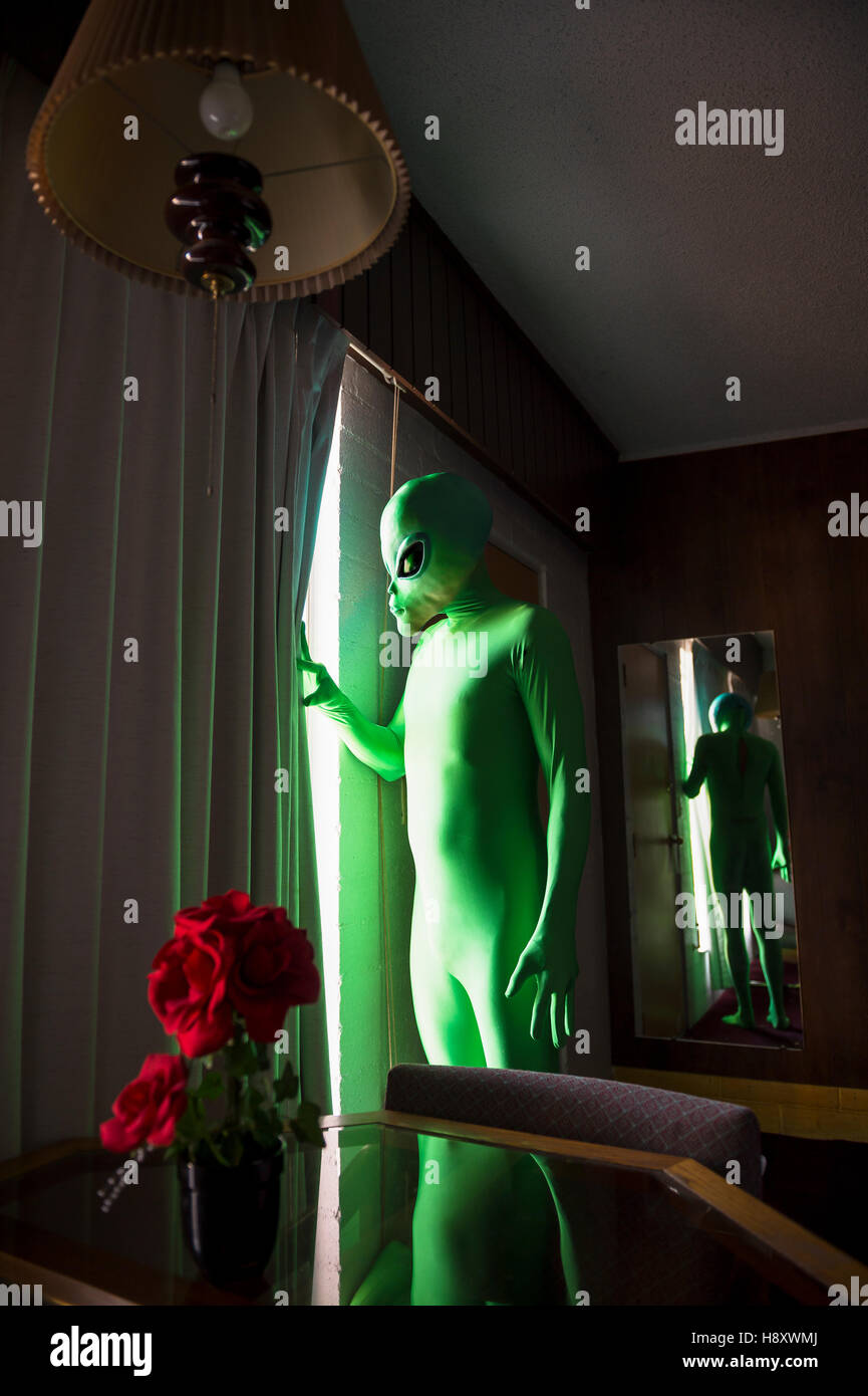 Green alien stands in a dark room pulling aside the curtains to look out the window Stock Photo