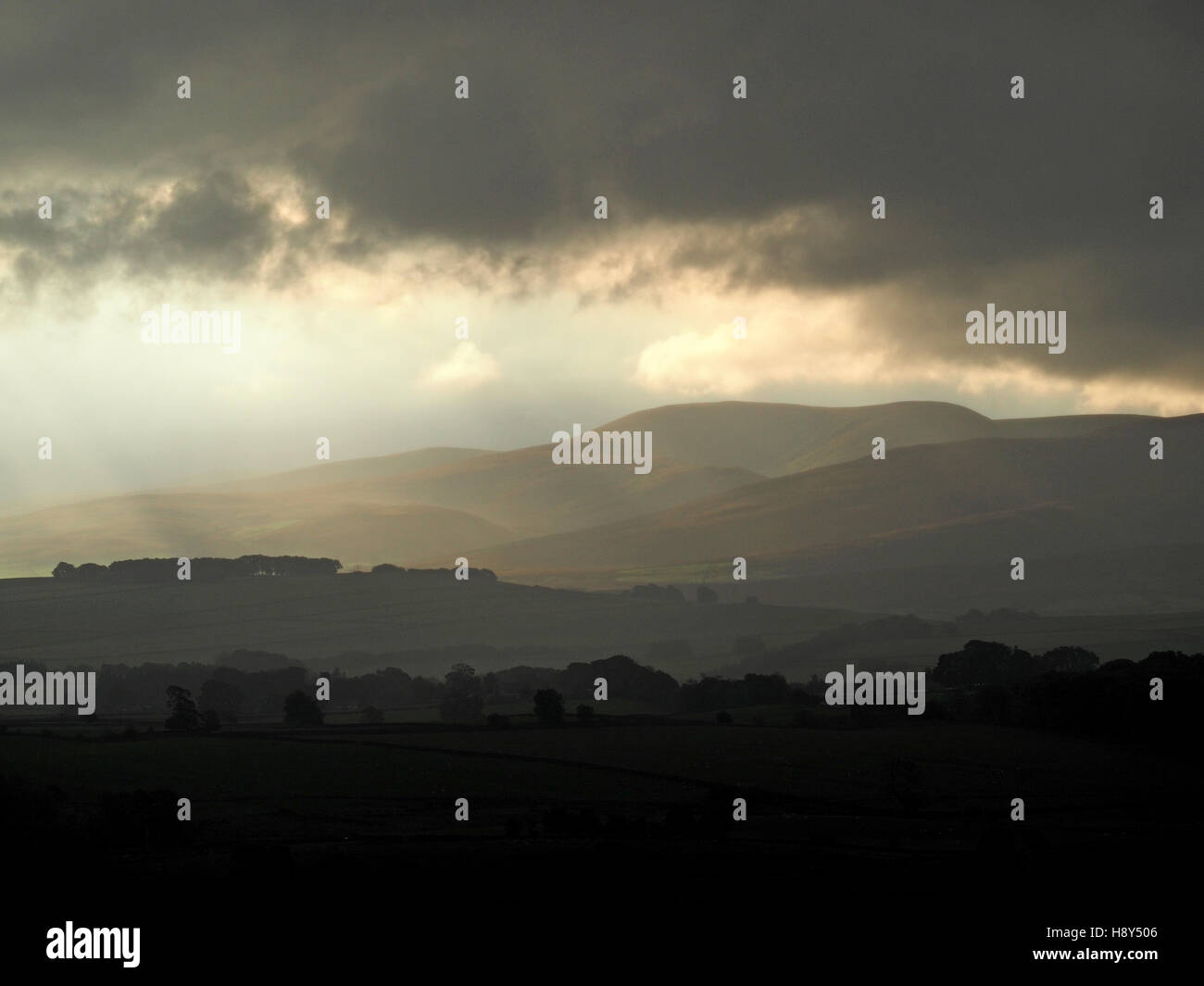 moody atmospheric golden morning light breaking through dark clouds onto receding foothills of Howgill Fells Cumbria England UK Stock Photo