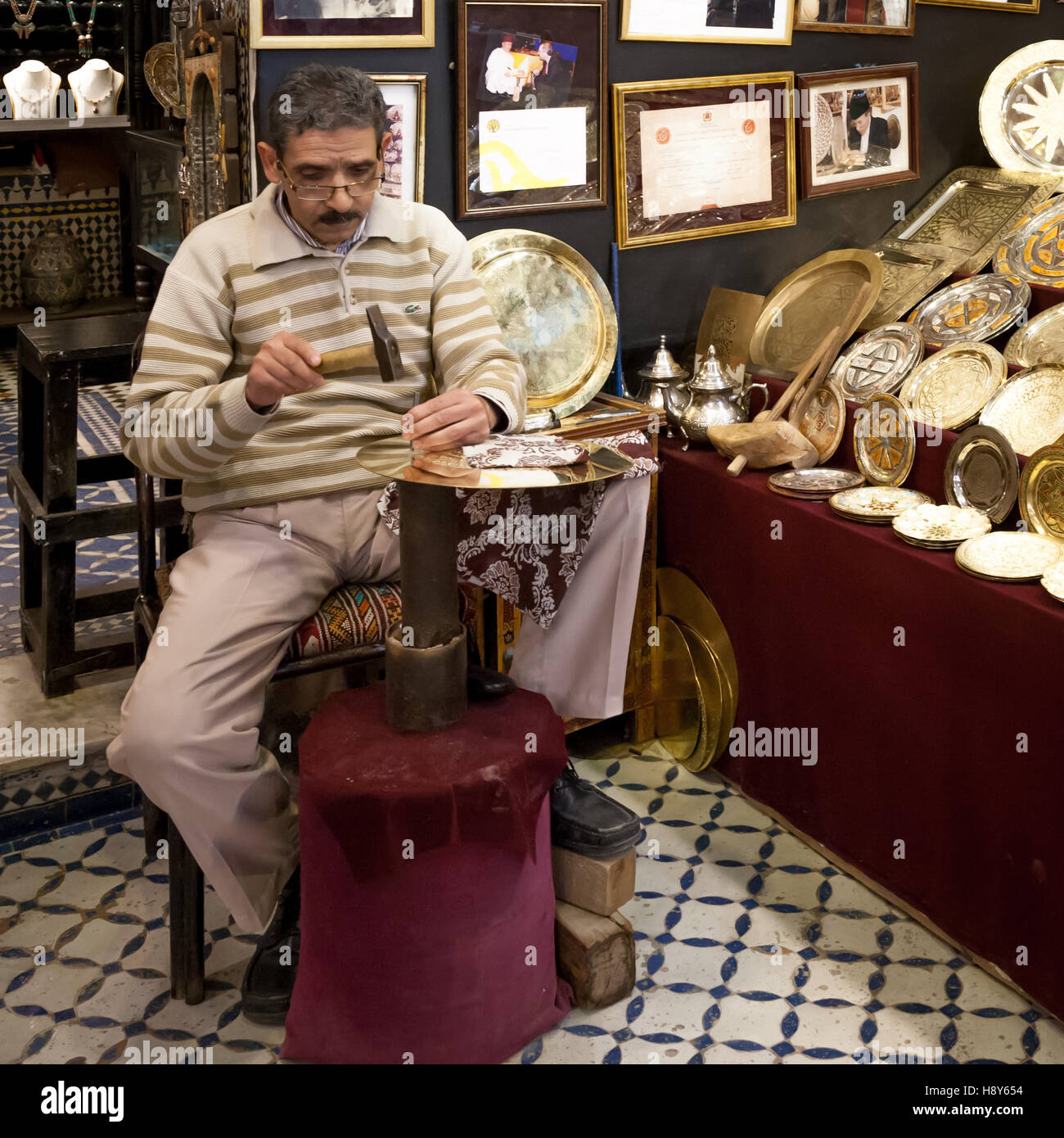 Brass craftsman Abdelgabar Guernani sits in his workshop in Fès, Morocco, engraving a brass plate with hammer and chisel. Numerous finished plates with different patterns are displayed in his shop, framed certificates hang on the wall. Stock Photo