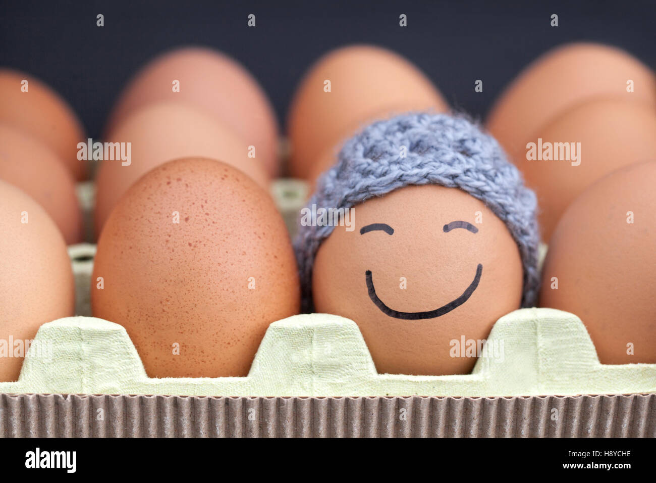 Smiling egg wearing a knitted hat souronded by blank brown eggs. Stock Photo