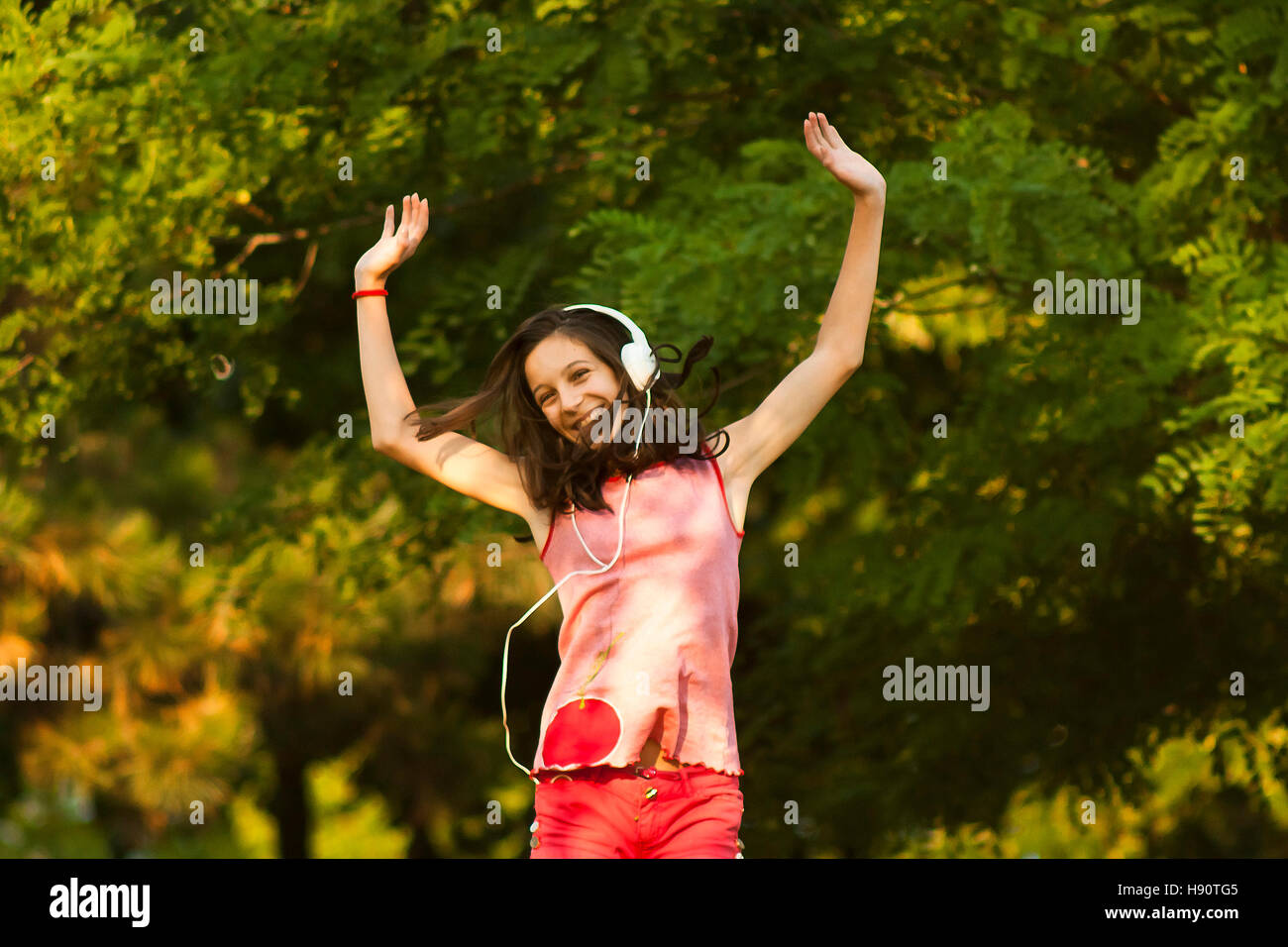 Happy teenage girl in red dancing in nature while listening to the music through headphones, smiling and looking at camera, on a sunny summer day. Stock Photo
