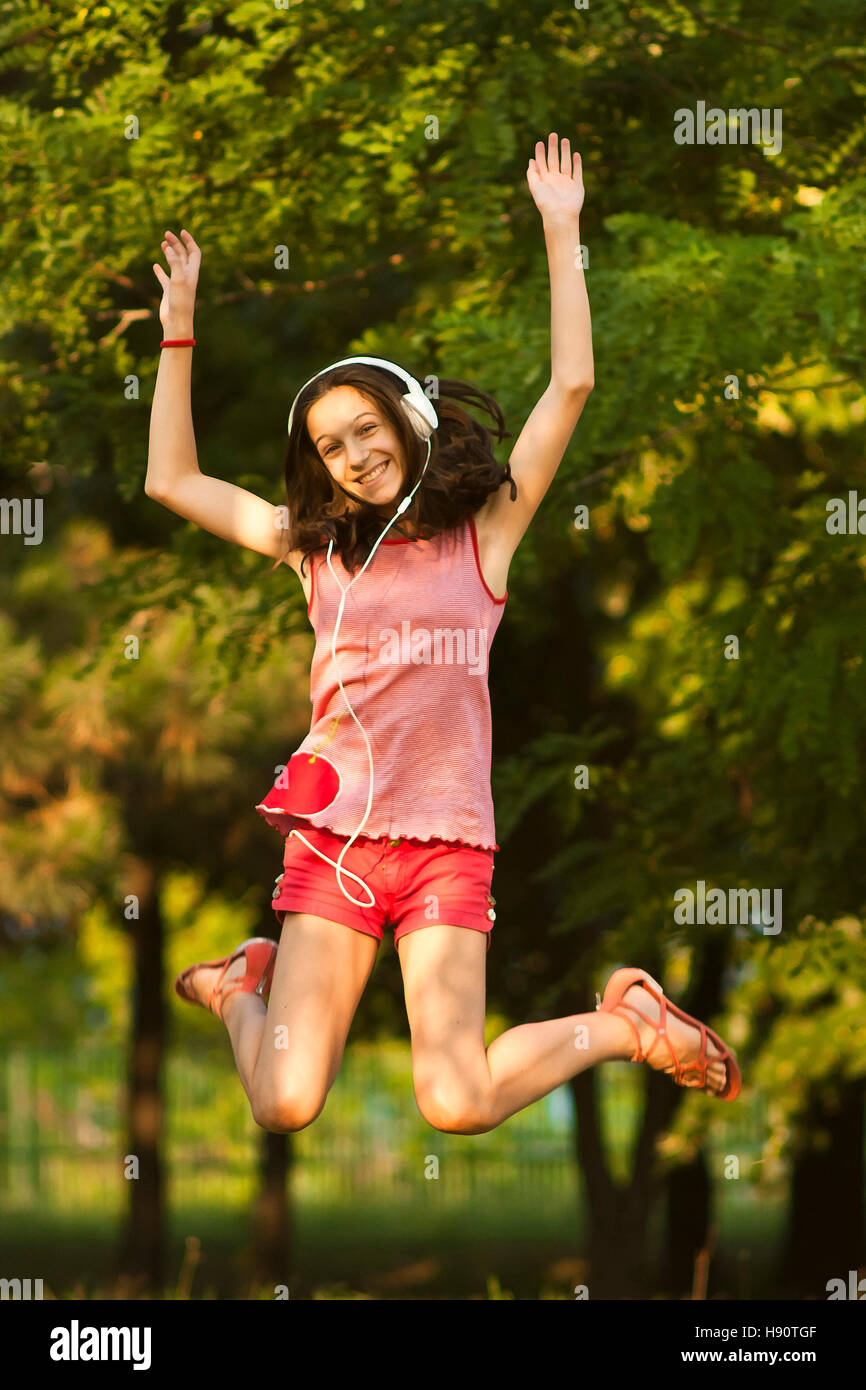 Happy teenage girl in red jumps in the park while listening to the music on headphones, smiling and looking at camera, on a sunny summer day. Stock Photo