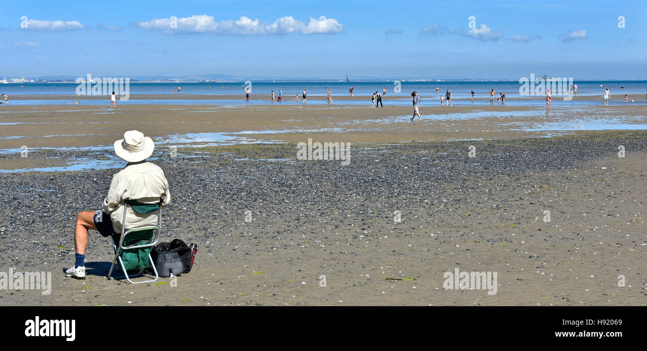 Older senior citizen man rear view on vacation sitting alone Ryde Isle of Wight England UK holiday beach low tide views of Solent people paddling sea Stock Photo