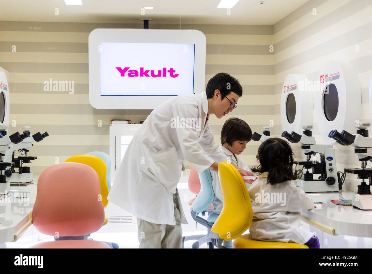 Japan, Nishinomiya, KidZania. 2 Children, boy, girl, in white laboratory coats using microscopes assisted by adult man in Yakult laboratory. Stock Photo