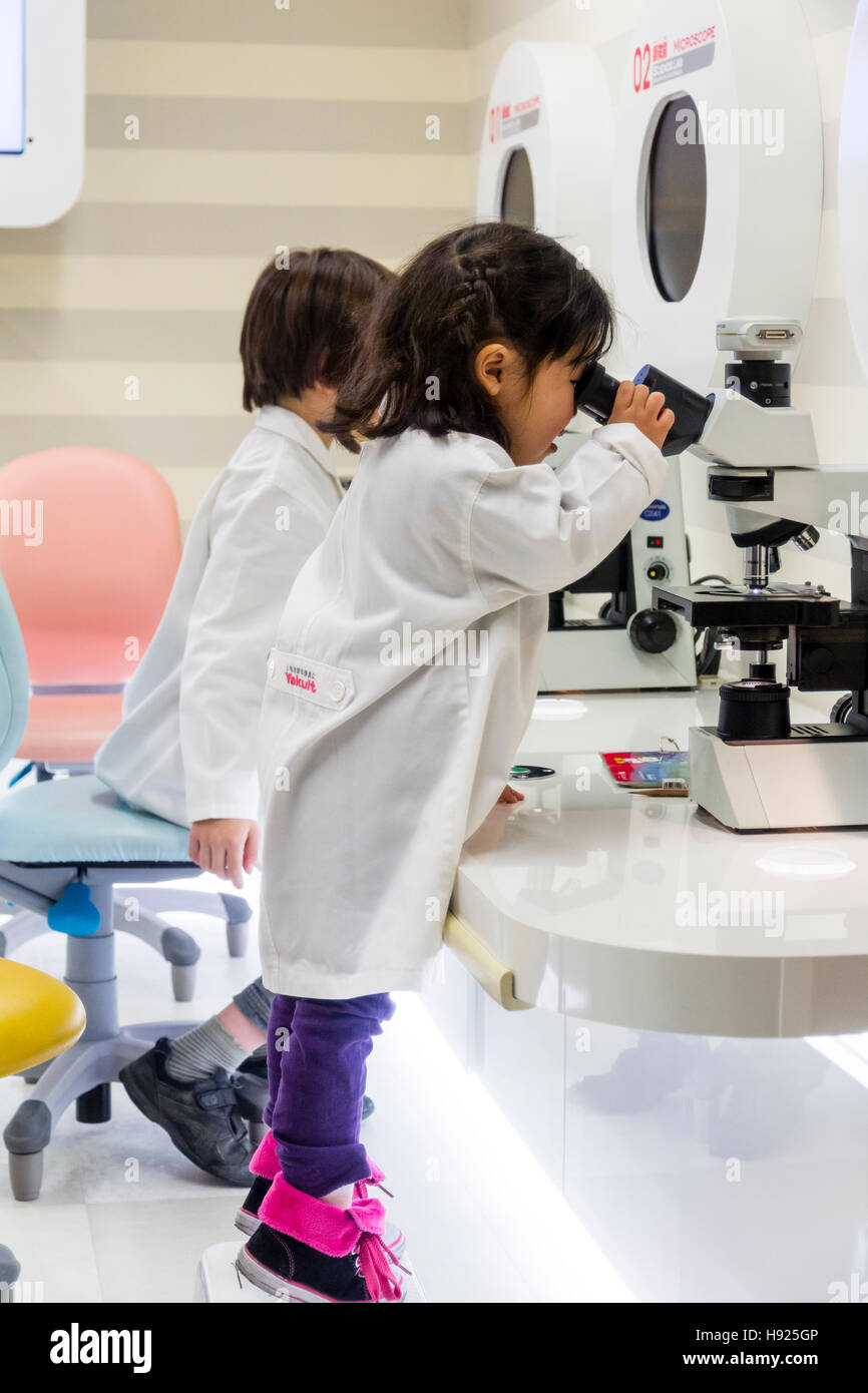 Japan, Nishinomiya, KidZania. Children, boy and girl in white laboratory coats using microscopes, little girl on tiptoe straining to view. Stock Photo