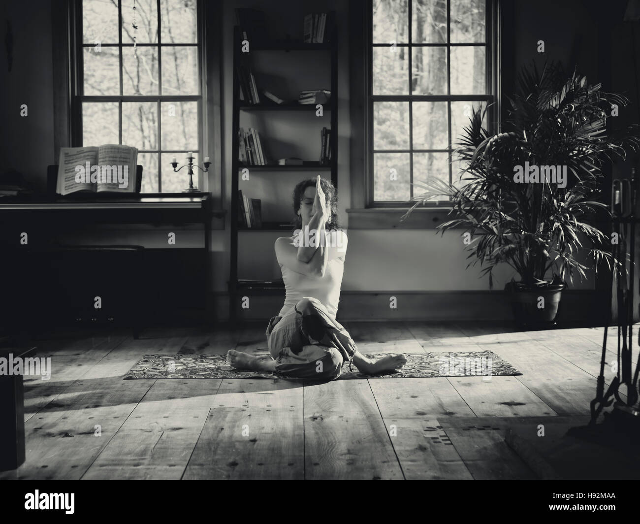 Artistic portrait of a woman sitting in a meditative yoga pose at home in a living room in dramatic light black and white Stock Photo