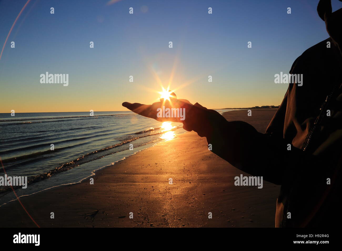 The silhouette of a man holds a seashell on the beach during sunset. A starburst or sunburst radiates from his hand. Stock Photo