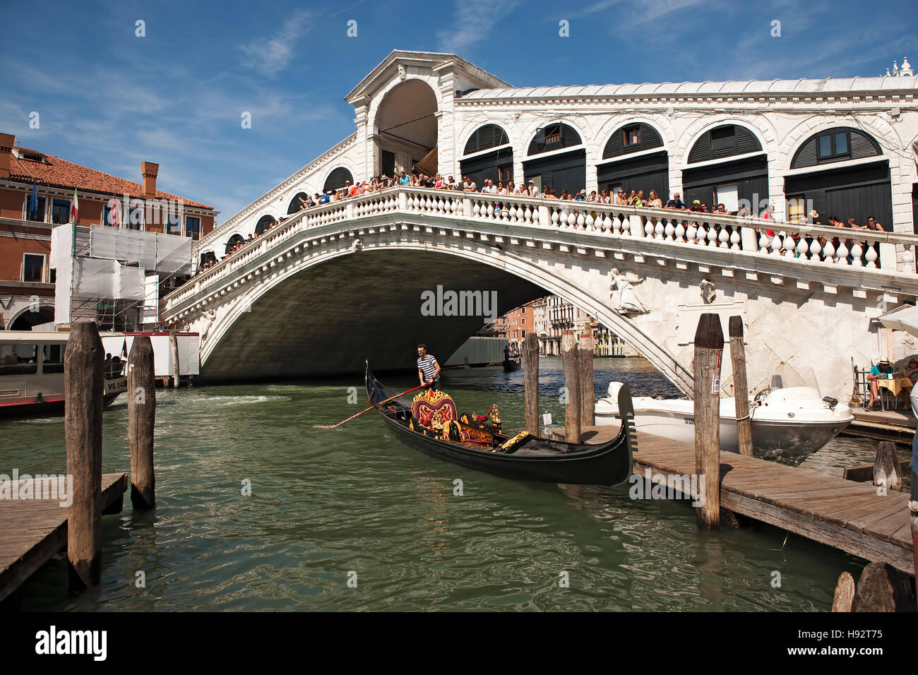 Crowds of people on the popular  Rialto Bridge watching tourists in a gondola by a wooden jetty, , Venice, Italy. Stock Photo