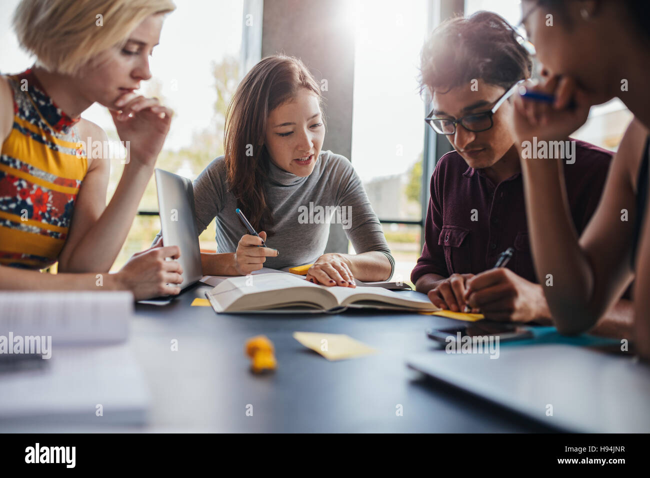 Multiethnic young people sitting at table reading reference books for study notes. Group of young students doing school assignment in library. Stock Photo
