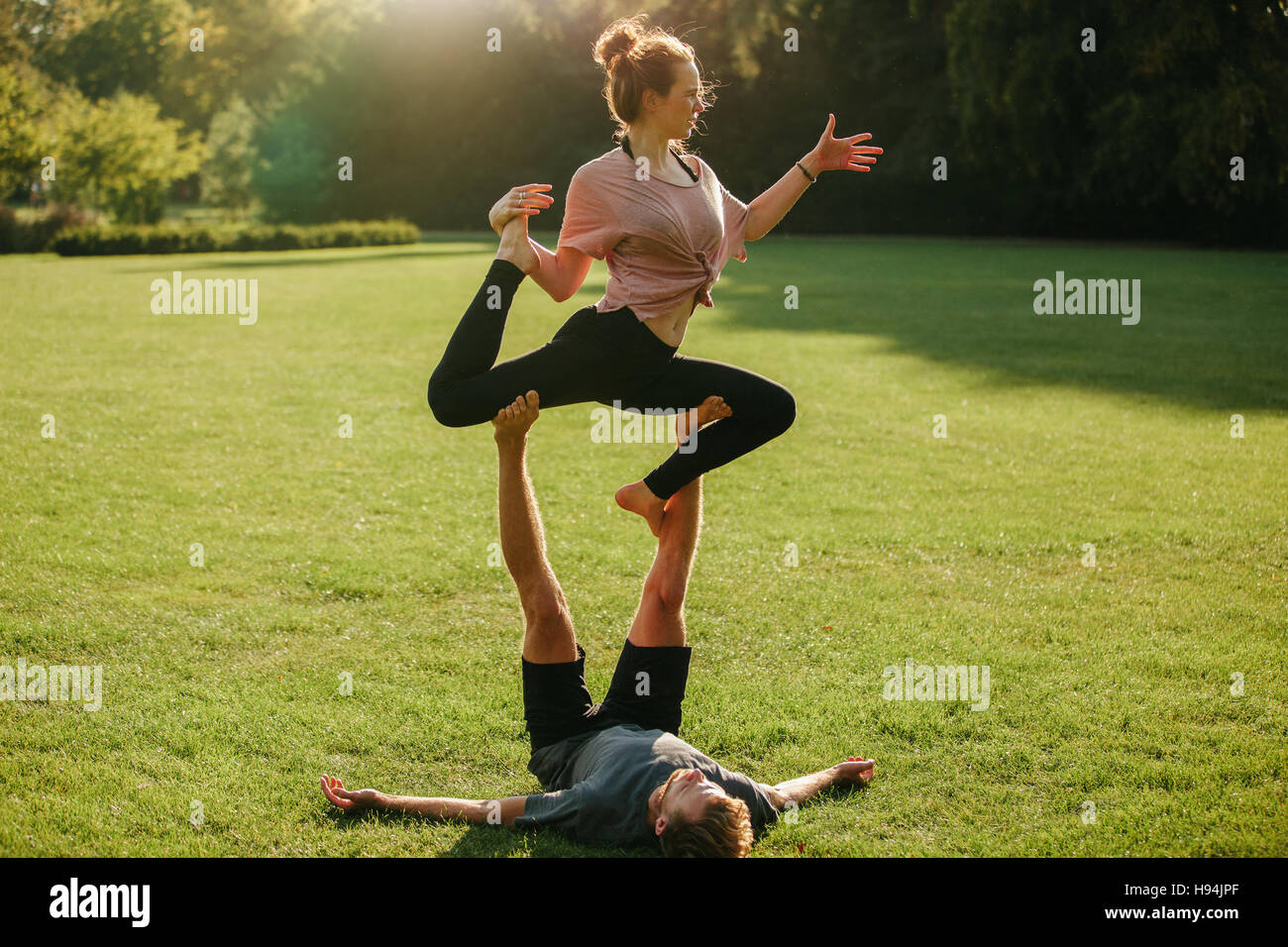 Man and woman doing various yoga poses in pair outdoors. Fit couple doing acro yoga in park, man balancing woman on his feet. Stock Photo