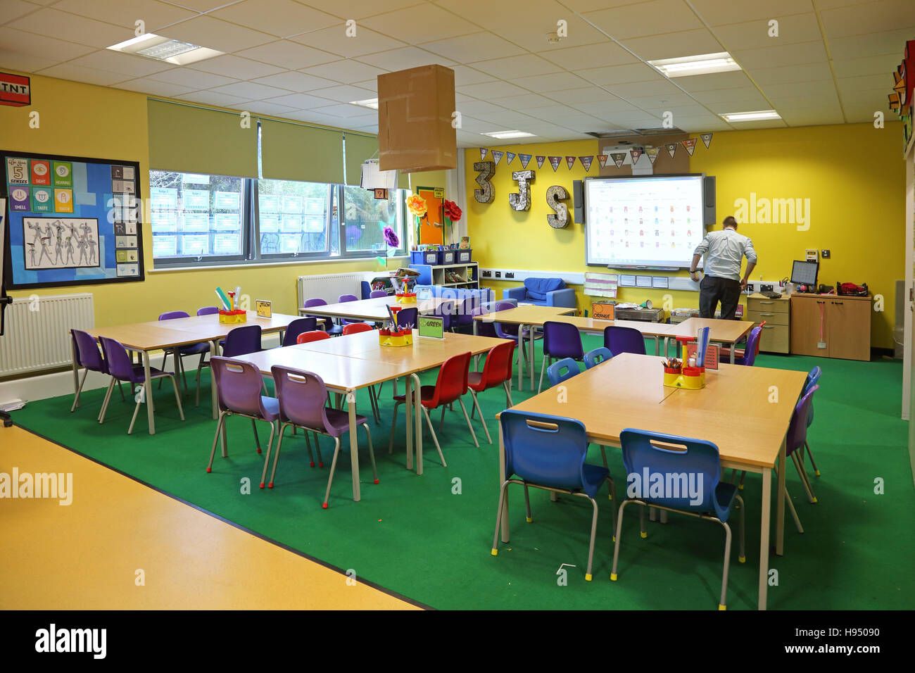 Brightly coloured interior of a modern year 1 school classroom showing tables, chairs and artwork on walls. Shows teacher. Stock Photo