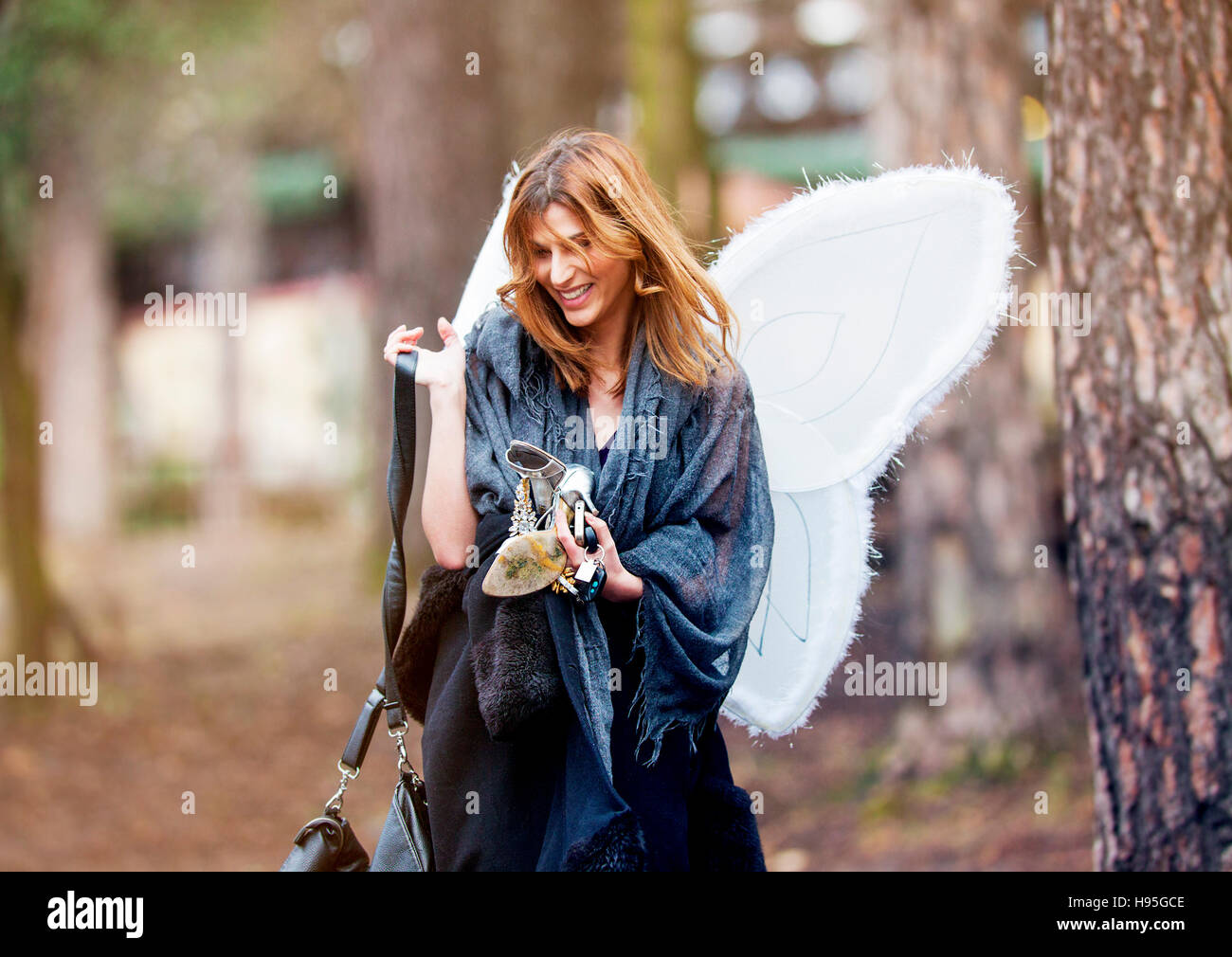 A beautiful happy smiling girl walking through the park with party angel wings and high heels in her hands. Stock Photo