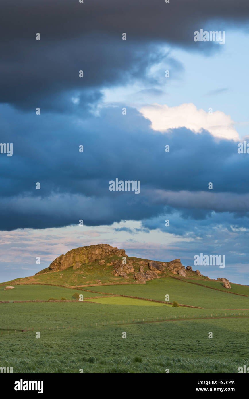 Under dark looming clouds & across green fields, a view of the distinctive rocky outcrop of Almscliffe Crag lit by sun - North Yorkshire, England, UK. Stock Photo