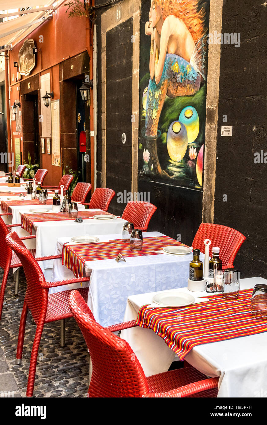 Colorful restaurant tables outside in an 'old town' street in Funchal, Madeira, with colourful mural on the wall Stock Photo