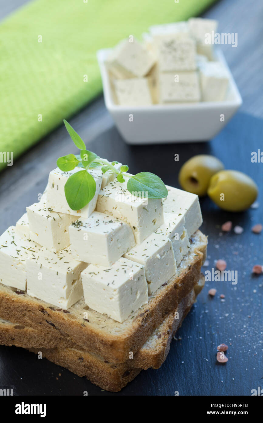 Tofu feta cheese with fresh oregano on a whole grain toast with linseed along with himalayan salt and two green olives on a black stone tray. Stock Photo