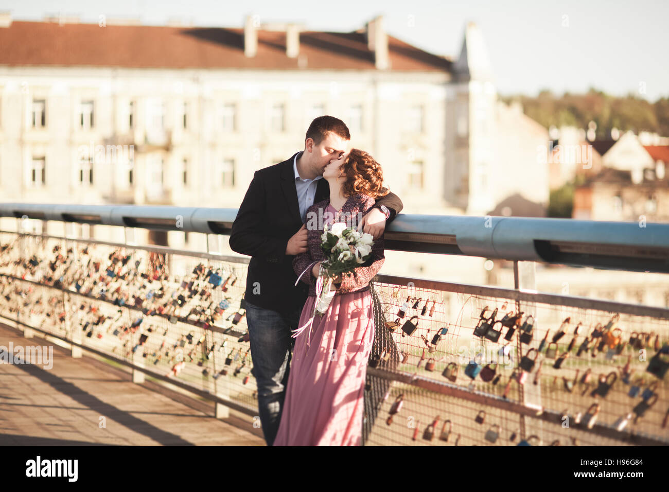 Stylish loving wedding couple, groom, bride with pink dress kissing and hugging on a bridge at sunset Stock Photo