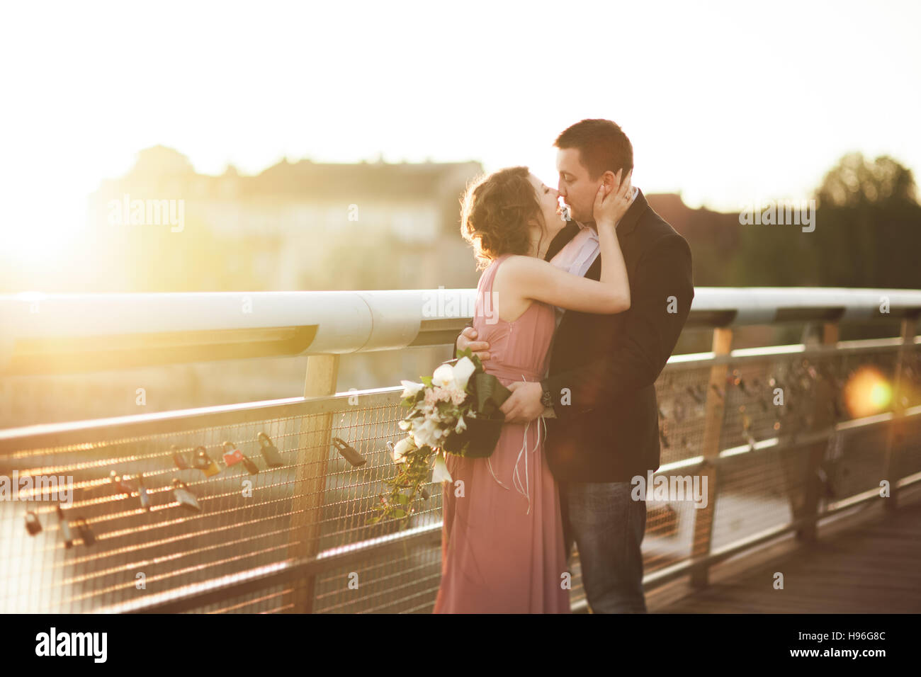 Stylish loving wedding couple, groom, bride with pink dress kissing and hugging on a bridge at sunset Stock Photo