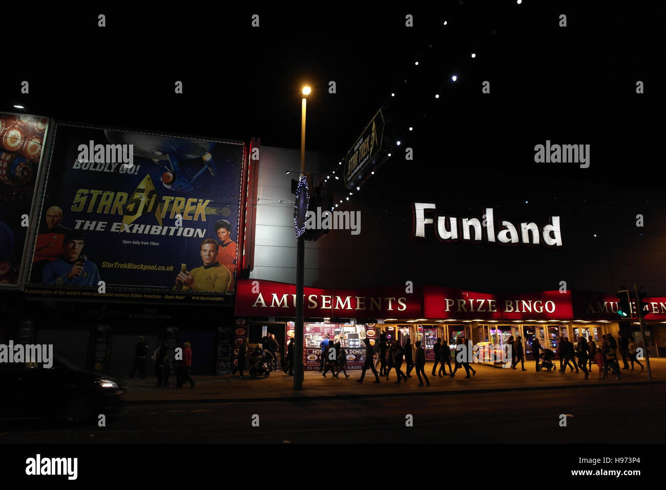 Night view across Central Promenade to people walking pavement front of Funland Amusement Arcade and Star Trek Illuminations, Blackpool, UK Stock Photo