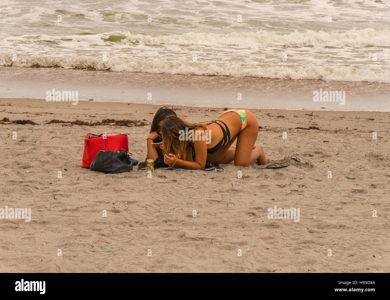 Two tanned girls lying or kneeling on the beach by the sea looking at their mobile phones Stock Photo