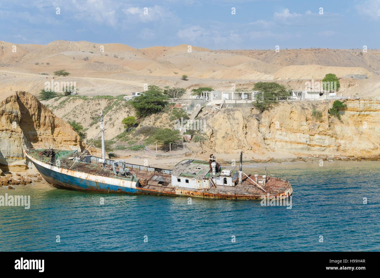 Rusting ship wreck stranded at beach of Angola's Namib coast near Caotinha. Concrete unfinished buildings and desert hills in background. Stock Photo