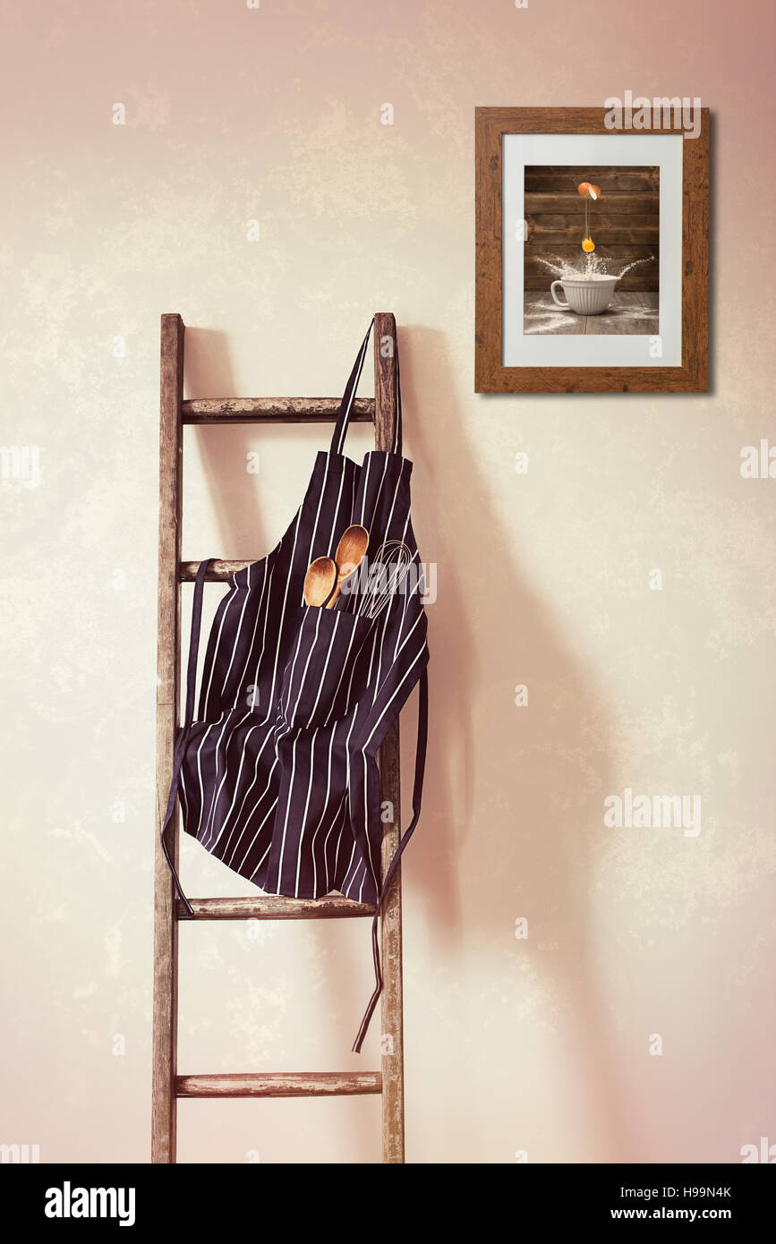 Kitchen apron hanging on the rung of a rustic wooden ladder with spoons and whisk Stock Photo